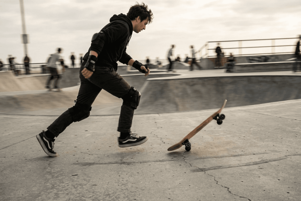 A skateboarder mid-bail stepping off the board onto concrete, wearing wrist guards and knee pads