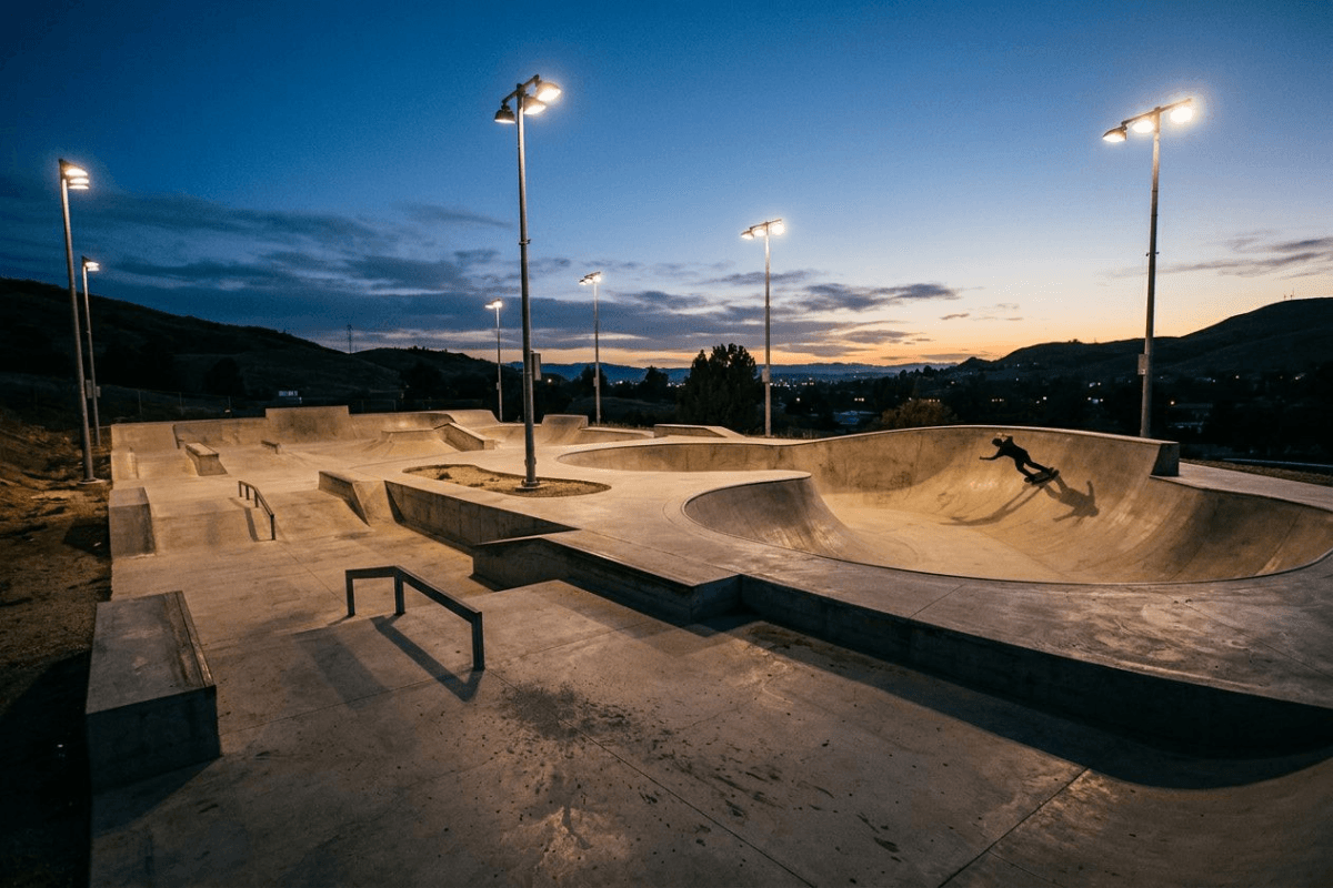 A concrete skate park at dusk lit by tall floodlights, a lone skater carving the bowl
